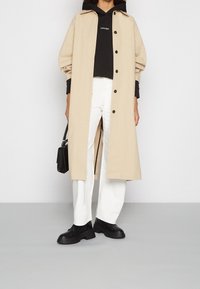 Persona che indossa un lungo cappotto beige sopra una felpa nera con cappuccio e pantaloni bianchi, tiene una piccola borsa nera e indossa stivali chunky neri.
