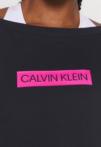 Tissu noir avec un large décolleté orné d'une étiquette rectangulaire rose vif portant l'inscription « CALVIN KLEIN » en lettres noires grasses.