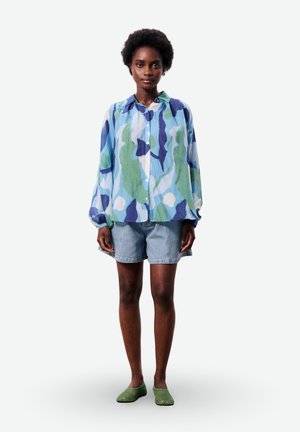 Femme debout portant un chemisier à manches longues bleu-vert à motifs, un short en jean clair et des chaussures slip-on vertes sur fond blanc.