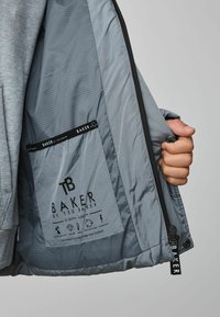 Veste grise avec un intérieur lisse et brillant affichant un motif à carreaux. Comprend une étiquette avec un logo, des instructions d'entretien et des icônes graphiques.