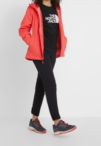 Rode waterdichte jas, zwart logo T-shirt, zwarte broek en grijze sportschoenen met rode accenten. Casual buitenoutfit geschikt voor activiteiten.