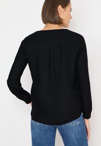 Blusa negra de manga larga con escote redondeado, tela suave y un detalle pliegue en la parte posterior. Combinada con vaqueros azules, mostrando un estilo casual.