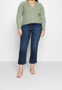 Cardigan côtelé vert clair à col en V avec fermeture à boutons, associé à un jean denim bleu foncé court et des sandales à talons hauts rose pâle.