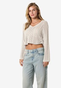 Krämfärgad, stickad kort tröja med V-ringning, långa ärmar och diamantmönster. Kombineras med ljusblå, högmidjade jeans.