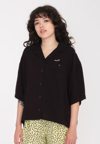 Chemise noire à boutons avec un col, des manches courtes et une poche avant. Présente un détail logo blanc, fabriquée en tissu léger.