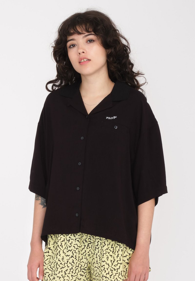 Chemise noire à boutons avec un col, des manches courtes et une poche avant. Présente un détail logo blanc, fabriquée en tissu léger.