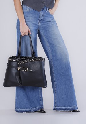Persona que lleva pantalones vaqueros azules de pierna ancha y zapatos negros puntiagudos, sosteniendo un gran bolso de cuero negro con detalles de hebilla y anillo.