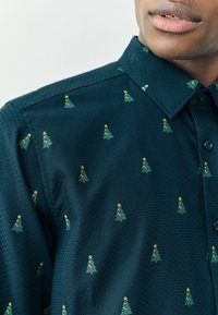 Mørkegrønblå skjorte med et tekstureret stof og et gentaget mønster af små juletræer, dekoreret med gule stjerner og røde accenter.