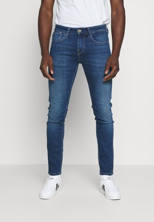 Homme portant un jean slim bleu foncé et des baskets blanches, debout devant un fond clair uni.