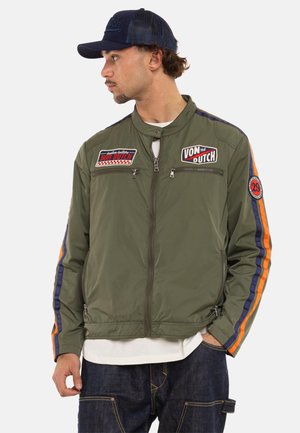 Homme portant une veste zippée vert olive avec des rayures orange et bleu marine sur les manches, plusieurs patchs, une casquette en maille bleu marine, une chemise blanche et un jean foncé.