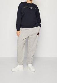 Felpa navy con logo bianco, abbinata a pantaloni della tuta grigi chiari con un emblema rosso. Il modello indossa sneakers bianche chunky.