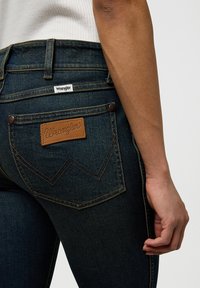 Dark blue denim jeans feature a leather patch with "Wrangler" logo and distinct back pocket stitching in a zigzag pattern.