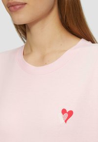 Roze katoenen t-shirt met een ronde hals en een kleine hartafbeelding in rood en wit op de linkerkant van de borst. Zachte textuur.