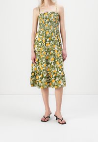 Robe d'été fleurie avec un corsage smocké, tissu vert orné de motifs d'agrumes jaunes, ourlet à volants et fines bretelles.