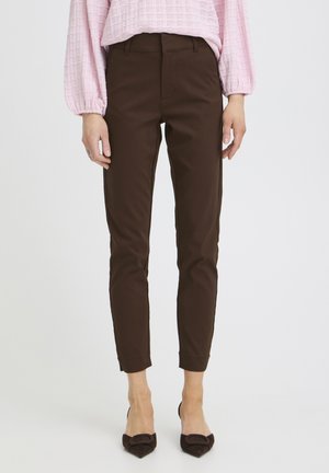 Frau trägt dunkelbraune, figurbetonte Hose, kombiniert mit einem rosafarbenen, strukturierten Langarmshirt und spitz zulaufenden braunen Schuhen.