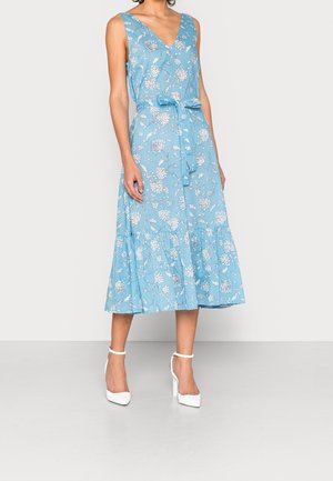 Femme portant une robe midi à fleurs bleue sans manches, avec un col en V et une ceinture à la taille, associée à des talons hauts blancs avec bride à la cheville.