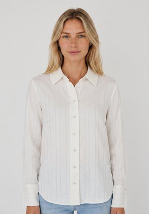 Femme aux longs cheveux blonds portant une chemise blanche à boutons à manches longues et un jean bleu clair, debout devant un fond uni.