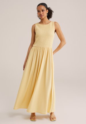 Femme portant une robe maxi jaune clair sans manches avec un corsage texturé et une jupe fluide, debout avec une main sur la hanche.