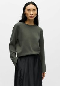 Oliwkowy sweter dzianinowy z okrągłym dekoltem i długimi rękawami, noszony na czarnej, zwiewnej spódnicy. Gładka tekstura, luźny krój.