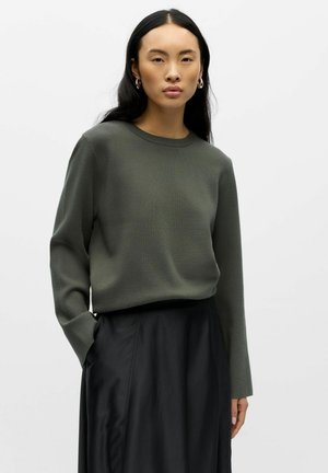 Oliwkowy sweter dzianinowy z okrągłym dekoltem i długimi rękawami, noszony na czarnej, zwiewnej spódnicy. Gładka tekstura, luźny krój.