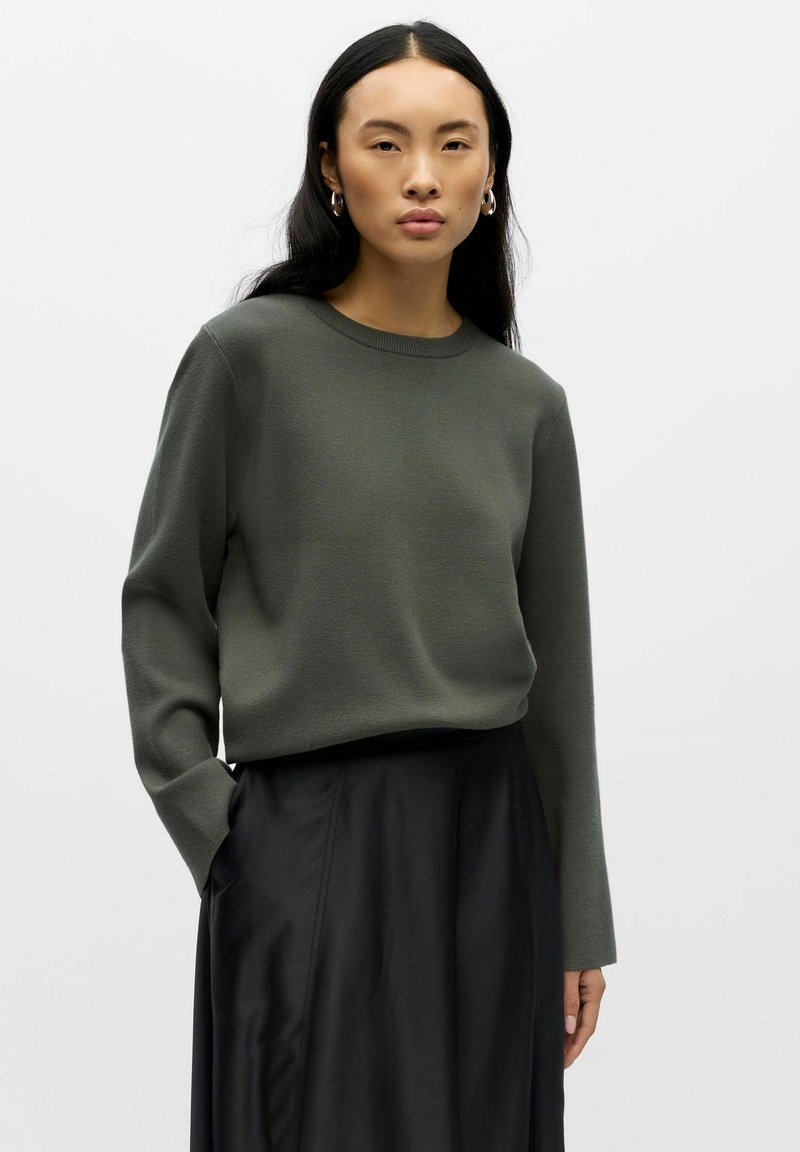 Oliwkowy sweter dzianinowy z okrągłym dekoltem i długimi rękawami, noszony na czarnej, zwiewnej spódnicy. Gładka tekstura, luźny krój.