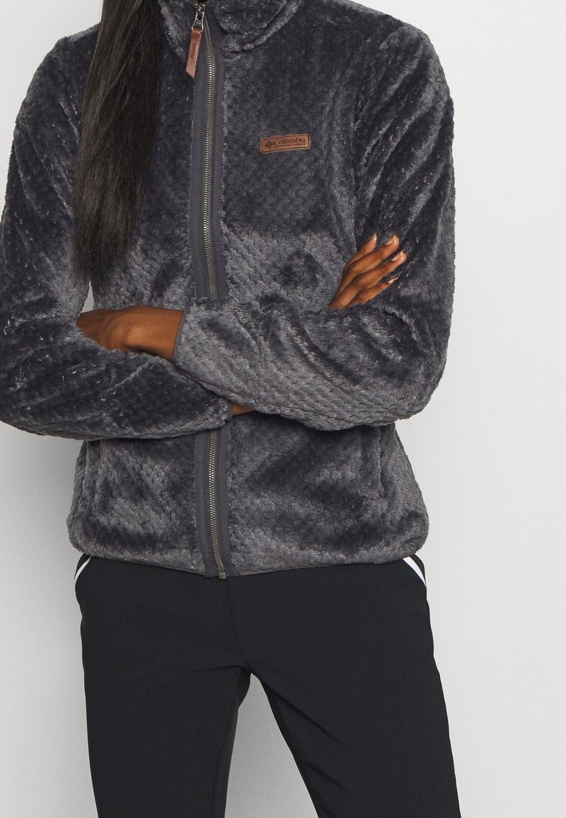 Grå fleece zip-up jacka med texturerat mönster, ståkrage och varumärkeslogotyp på bröstet. Kombinerad med svarta träningsbyxor.