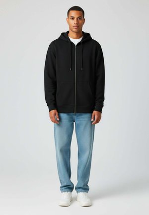 Zwarte zip-up hoodie met trekkoorden, voorzakken, gedragen met lichtblauwe spijkerbroeken en witte sneakers. Smooth materiaal en relaxte pasvorm.