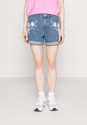 Person iført blå denimshorts med hvidt blomsterbroderi, lyserød top, hvide sokker og lyserøde og hvide sneakers, stående mod en ensfarvet baggrund.