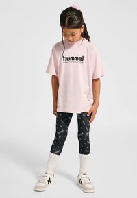 Ljusrosa Hummel-t-shirt, svarta leggings med vita mönster, vita strumpor och ljusa sneakers med svarta detaljer, stående position.