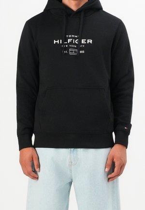 Persona que lleva una sudadera con capucha negra de Tommy Hilfiger con texto del logo en blanco y vaqueros azul claro sobre un fondo claro y liso.