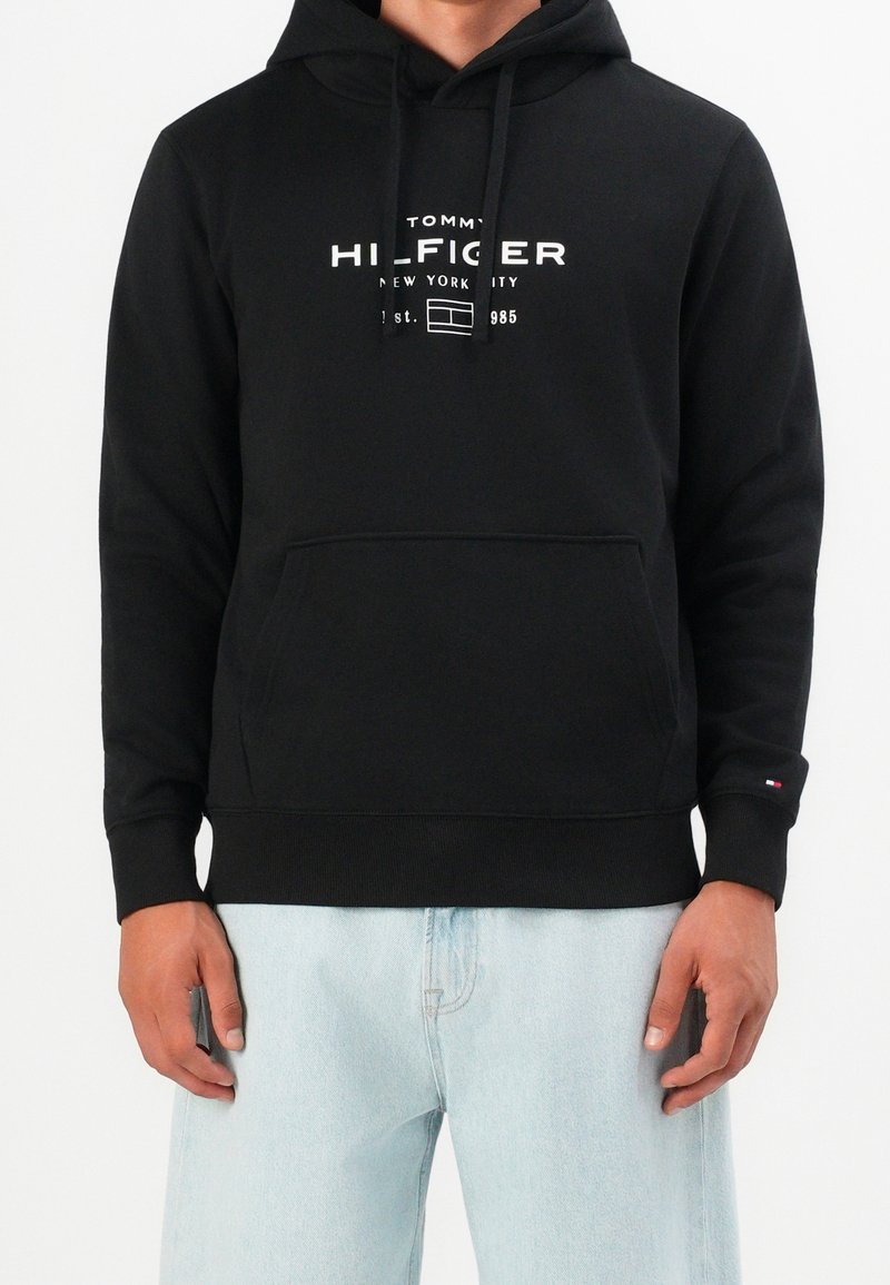 Personne portant un sweat à capuche noir Tommy Hilfiger avec un logo blanc et un jean bleu clair devant un fond uni clair.