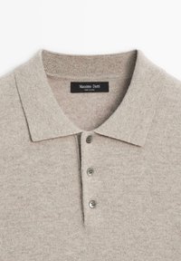 Beige stickad polo med krage, tre grå knappar och en etikett som säger "Massimo Dutti." Strukturerad tyg med en mjuk känsla.