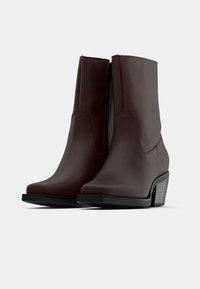 Par de botas de cuero marrón oscuro hasta la mitad de la pantorrilla, con cremalleras laterales y tacones de bloque, sobre un fondo claro y liso.