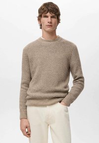 Maglione beige lavorato a maglia con motivo a spina di pesce, scollo rotondo e polsini e orlo a coste, indossato con pantaloni chiari.