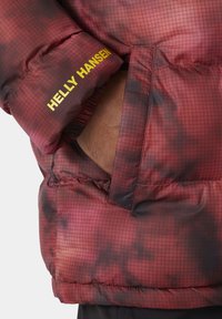 Přepravní bunda s červeným a černým kostkovaným vzorem a elastickými manžetami. Obsahuje viditelnou kapsu a žluté logo "Helly Hansen" vyšité na rukávu.