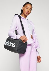 Juodas sportinis krepšys su baltu „adidas“ logotipu, pagamintas iš glotnaus audinio su tinklinėmis detalėmis, nešamas su paminkštintu peties diržu.