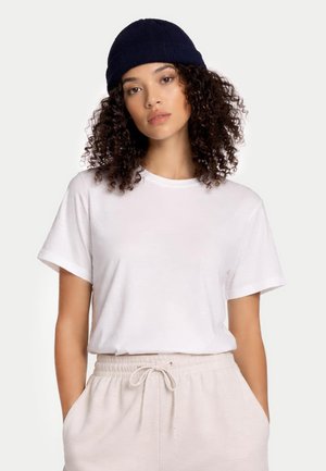 Junge Frau mit lockigem Haar, die eine marineblaue Mütze, ein weißes T-Shirt und hellbeige, mit Kordelzug versehene Hose trägt, steht mit den Händen in den Taschen.