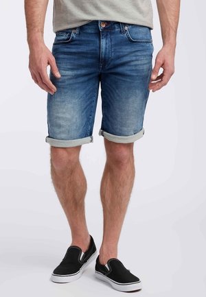 Homme portant un short en denim bleu retroussé, un t-shirt gris et des chaussures noires sans lacets, debout devant un fond clair uni.