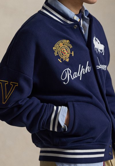 Ναυτικό μπλε μπουφάν varsity με λευκές ρίγες, κεντημένα σχέδια και το γράμμα "V" στο μανίκι. Φοράει ένα ανοιχτό μπλε πουκάμισο με γιακά από κάτω.