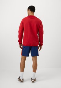 Κόκκινη μπλούζα με στρογγυλή λαιμόκοψη, μακριά μανίκια, συνδυασμένη με μπλε σορτς με πλαϊνές ρίγες. Μοντέλο που στέκεται με την πλάτη προς την κάμερα σε απλό φόντο.