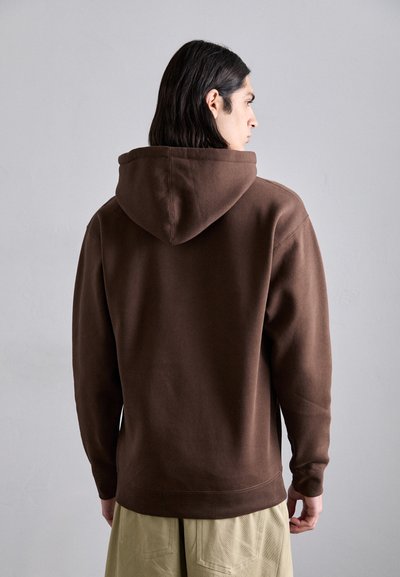 Brown cotton hoodie with a spacious hood, relaxed fit, and ribbed cuffs. Visible stitching detail along the hem. Worn with beige shorts.