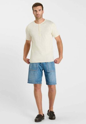 Man met een beige henley-shirt met korte mouwen, blauw denim korte broek tot aan de knieën met steekzakken aan de voorkant, en zwarte veterschoenen, staand tegen een witte achtergrond.