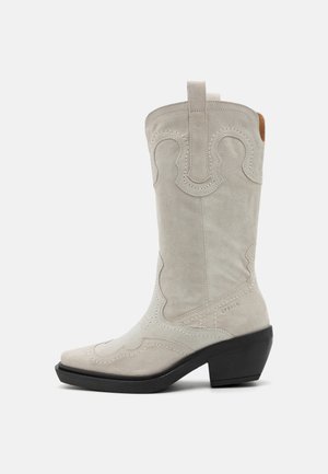 Cizme cowboy din suede alb, cu vârf ascuțit, cusături decorative și un toc mic pătrat. Include un tab pentru tragere în partea de sus pentru o îmbrăcare ușoară.