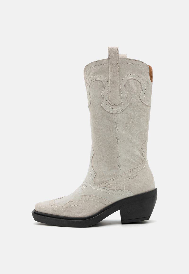Bota de cowboy em camurça branca com bico pontudo, costura decorativa e um salto bloco curto. Apresenta uma alça na parte superior para facilitar o calce.