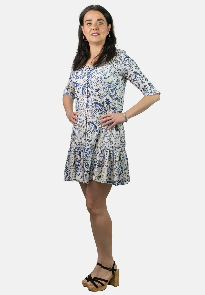 Vestido con estampado floral en azul y blanco, con cierre de botones en el frente, mangas de tres cuartos y dobladillo con volantes en capas. Combinado con sandalias plataforma gruesas.