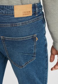 Temno modre jeans hlače iz jeansa z usnjenim ovratnikom na pasu, na katerem je napis "DENIM POTOVANJE". Imajo en zadnji žep s kontrastnim šivanjem.