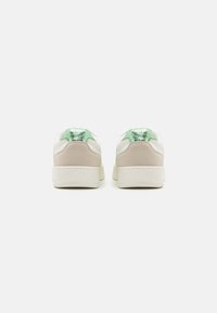 Baskets blanches et beiges avec un accent métallique vert au talon, des panneaux en suède texturé et une finition lisse. Forme arrondie avec un profil bas.