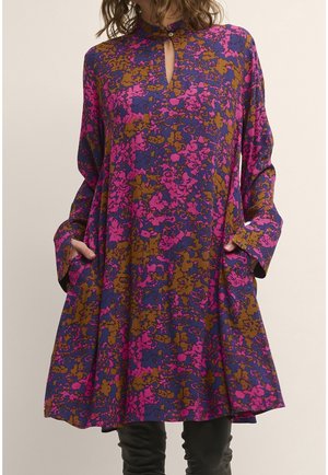 Kvinde iført en løs, langærmet kjole med pink, lilla og brun abstrakt blomsterprint og forlommer.