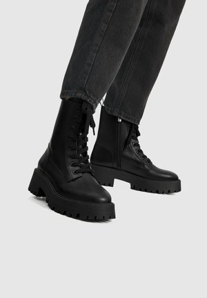 Sorte snørestøvler med kraftige såler, båret med mørke denimjeans mod en neutral baggrund.