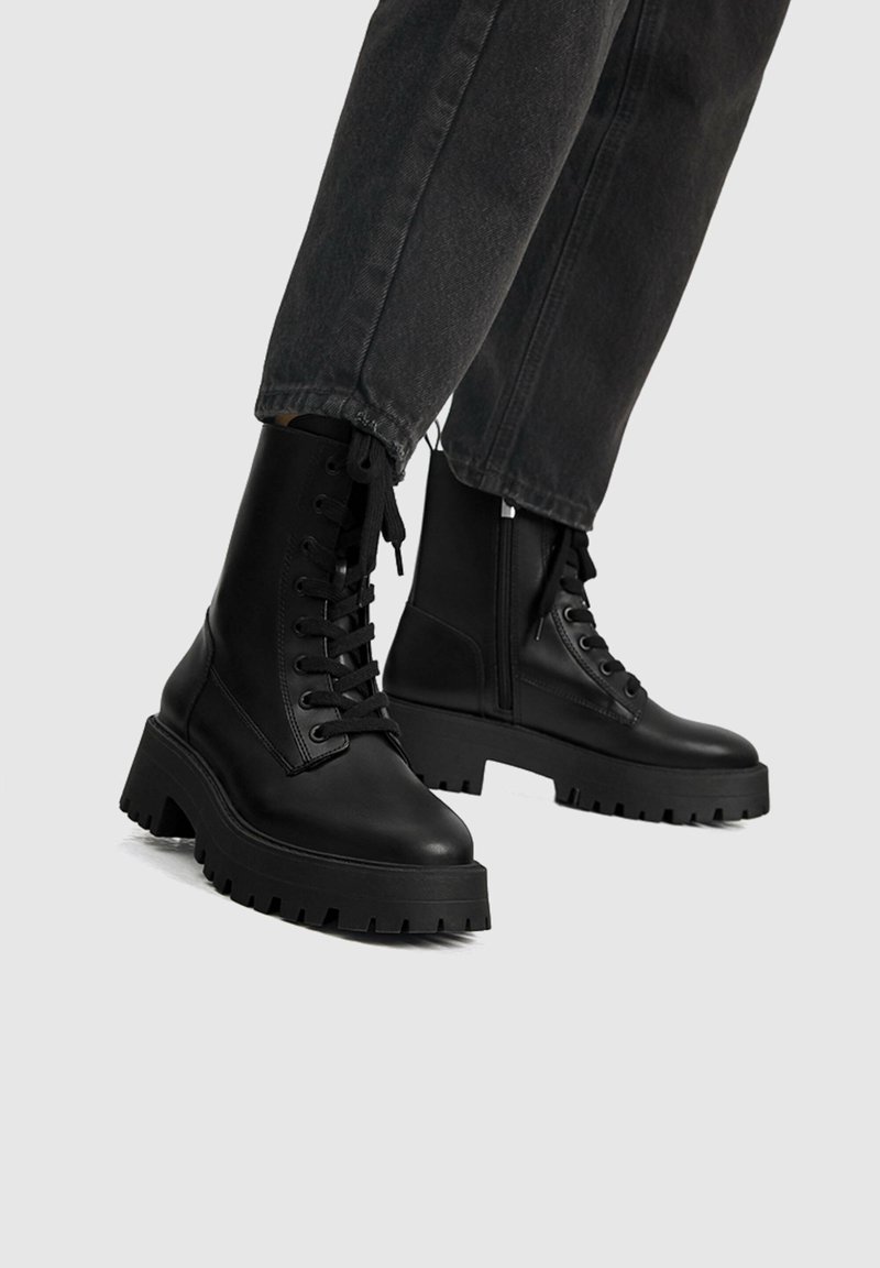 Bottes de combat noires à lacets avec semelles épaisses portées avec un jean en denim foncé sur un fond neutre.